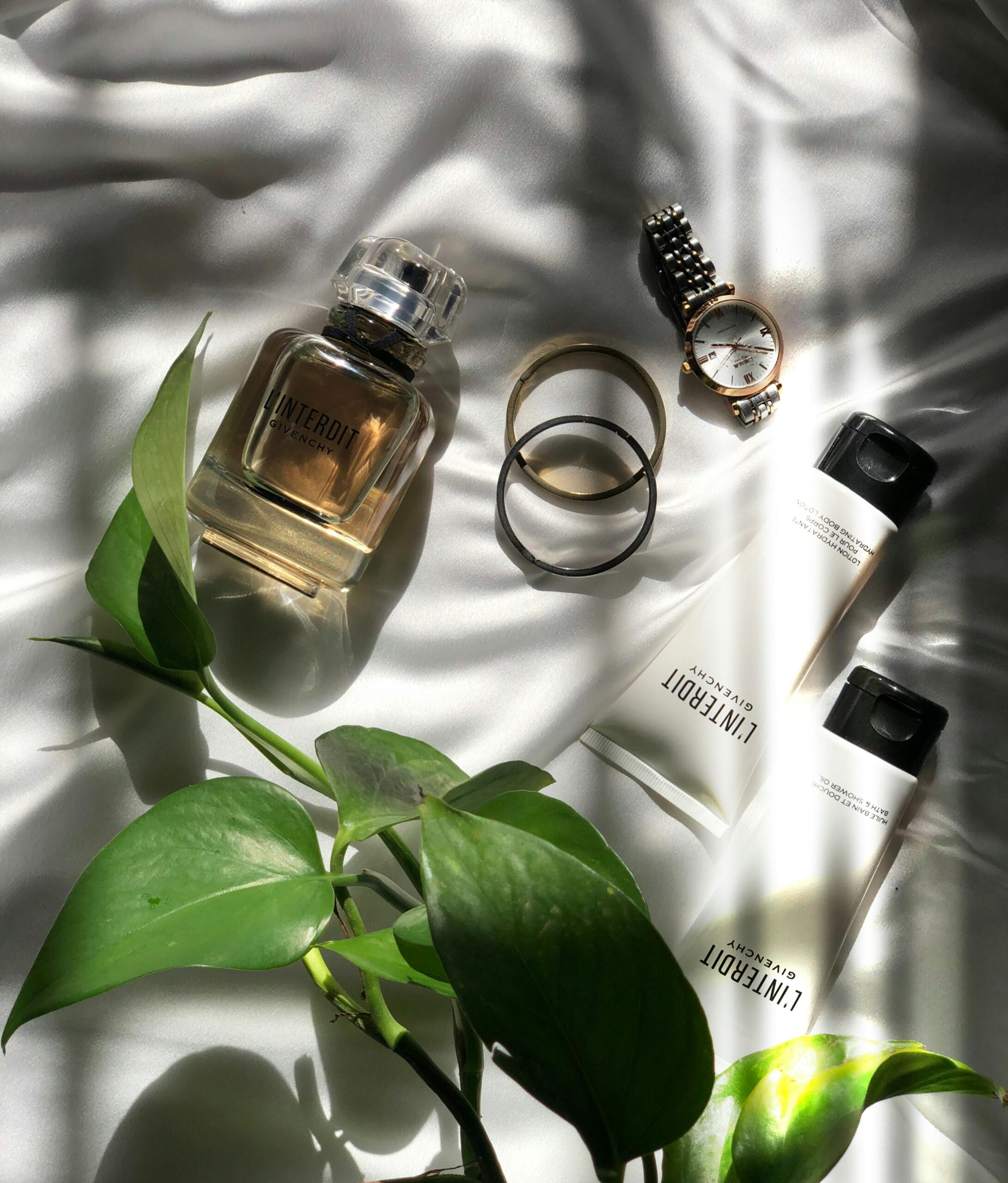 Flat lay of perfume, cosmetics, watch, and rings on a white surface with sunlight and green leaves.